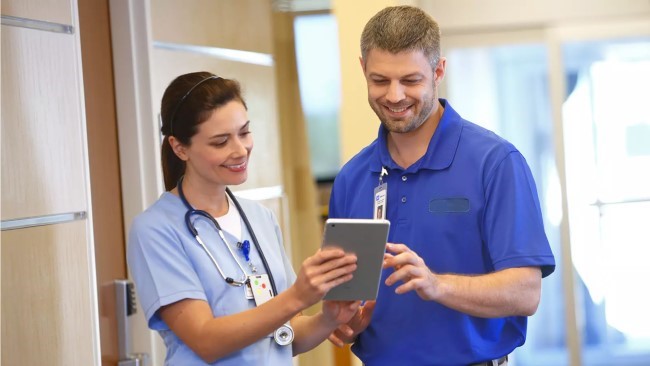 A clinician and a Hillrom service technician smile while looking at a tablet in a hospital hallway